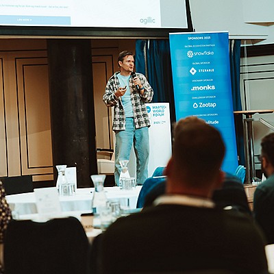 "Speaker presenting on stage at with sponsor banner and audience in view.