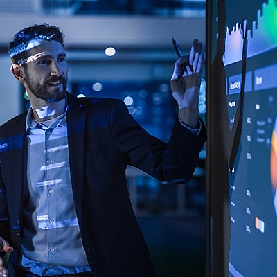 A man in a suit presents data on a large digital screen, gesturing with a pen. 
