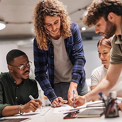 Four colleagues collaborating around a table, focused on documents. The setting is casual and engaged, conveying teamwork and creativity.