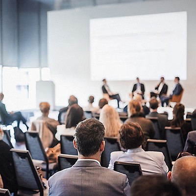 Audience attending a conference panel in a modern, bright room. Four speakers sit onstage, engaging in discussion.
