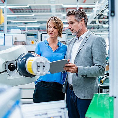 Two professionals in a high-tech manufacturing setting discuss a robotic arm.