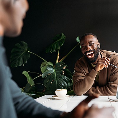 A man in a brown sweater laughs joyfully, holding glasses in a cafe. 