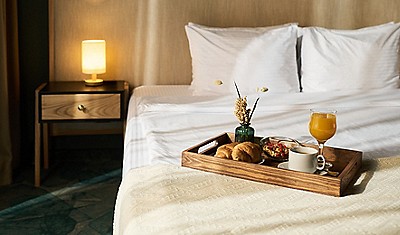 Cozy hotel room showcased with breakfast tray on bed. Tray filled with coffee, juice, pastries, and fruit, with warm lamp providing ambient lighting next to bed