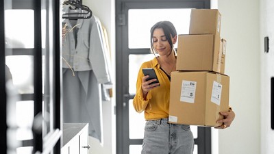 A woman in a yellow shirt holds three cardboard boxes while checking her phone. She stands by a doorway, looking content and focused.