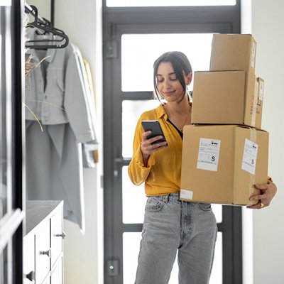 A woman in a yellow shirt holds three cardboard boxes while checking her phone. She stands by a doorway, looking content and focused.