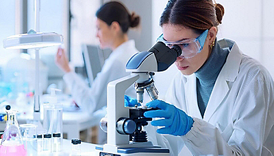 Two women in a laboratory. One woman with goggles is looking into a microscope