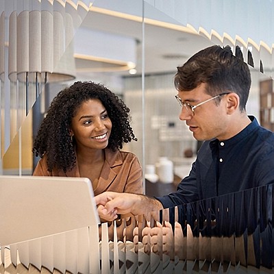 A woman and man interact positively at a laptop in a modern office. They are engaged and smiling, suggesting a collaborative and friendly atmosphere.