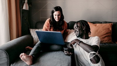 Two people sit on a couch in a living room; one uses a blue laptop, the other holds a smartphone. Their faces are obscured. The room features grey walls, an orange pillow, and a floor lamp in the background.