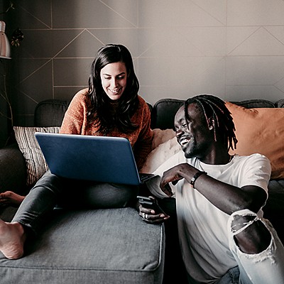 Two people sit on a couch in a living room; one uses a blue laptop, the other holds a smartphone. Their faces are obscured. The room features grey walls, an orange pillow, and a floor lamp in the background.