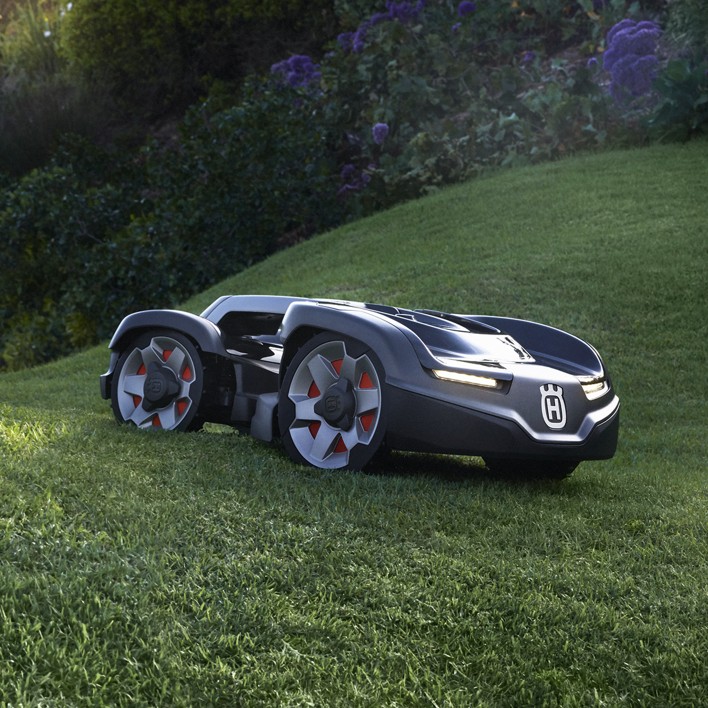 Robotic Husqvarna lawn mower on a grassy slope with bushes and purple flowers in the background.