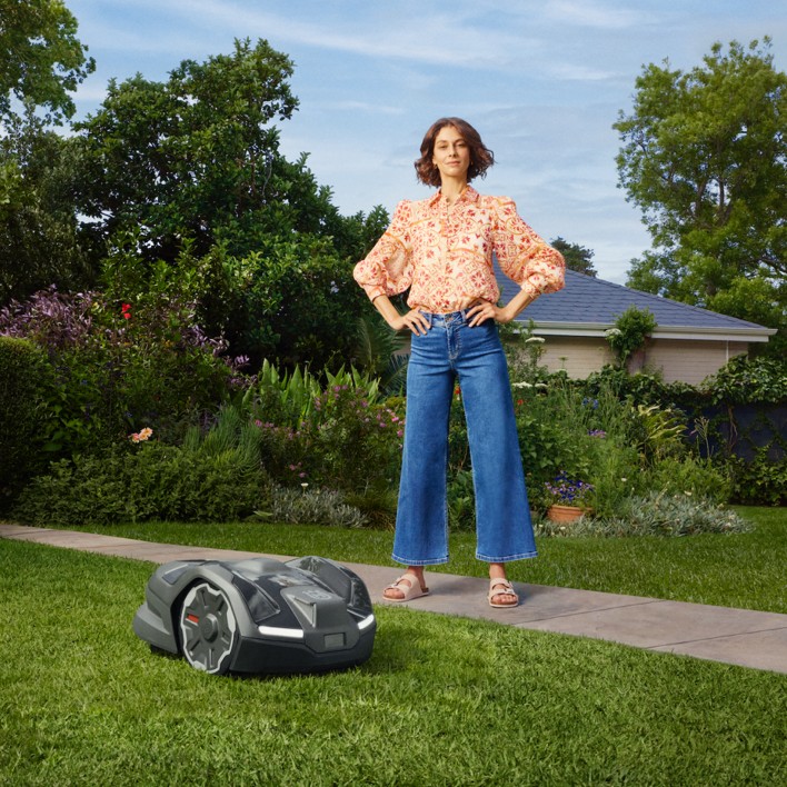Person standing on a lawn with a robotic mower in front, garden and house in the background.