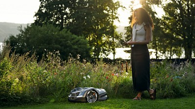 Person holding a device in a garden with tall grass, watching a Husqvarna lawn mower.