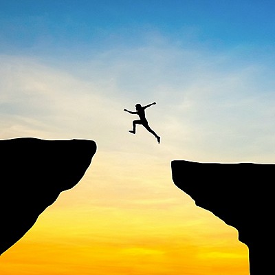 Silhouette of person jumping between two cliffs at sunset.