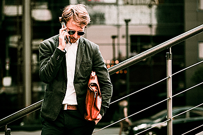 The Young Man Is Standing On The Stairs In The City Center. He Is Holding A Brown Briefcase Under His Arm and Speaking On His Phone. He Is Looking Down. Toned image.