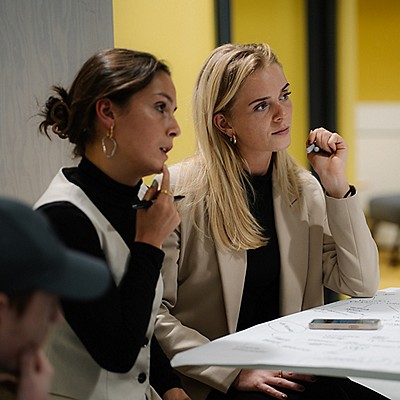 Two people sit at a table, holding pens and reviewing a document, with a yellow background and a third person partially visible on the left.