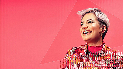 Person with short blonde hair smiling against a bright pink background, wearing a colorful patterned jacket.