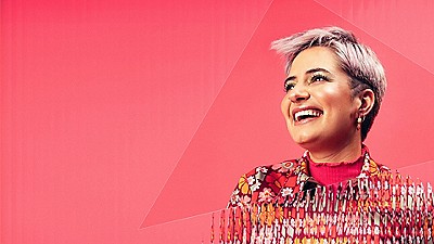 Person with short blonde hair smiling against a bright pink background, wearing a colorful patterned jacket.