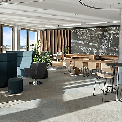 Moderne Office-Lounge mit großen Fenstern, viel Tageslicht, Holztischen, hellen Stühlen, Zimmerpflanzen und einer großen Wandgrafik, gestaltet als offener, kollaborativer Arbeitsbereich.