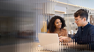 A woman and man interact positively at a laptop in a modern office. They are engaged and smiling, suggesting a collaborative and friendly atmosphere.
