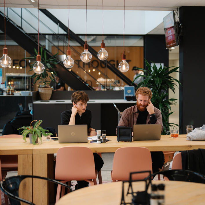 Two of our colleagues sitting in the canteen, working behind their laptop