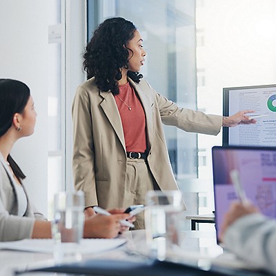 A woman in a beige suit presents data on a screen to colleagues in a modern office. She points to a pie chart.