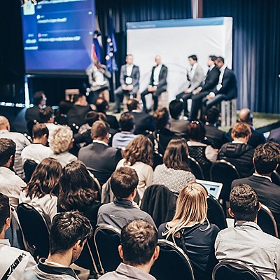Audience watches a panel discussion in a conference hall. Five people on stage, with a large screen displaying text.