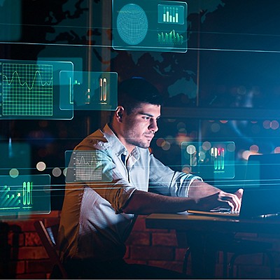 A man works intently on a laptop at night, surrounded by virtual data graphs and charts.