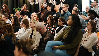 Group of people seated in a room with hanging lights, listening attentively.