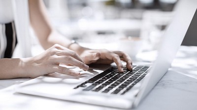 A close-up of hands typing on a laptop keyboard, set against a bright, airy cafe background with lightly blurred details.