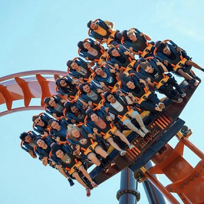 A roller coaster car full of excited riders, strapped in with orange harnesses, tilts downward against a clear blue sky.