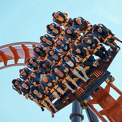 A roller coaster car full of excited riders, strapped in with orange harnesses, tilts downward against a clear blue sky.