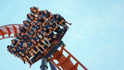 A roller coaster car full of excited riders, strapped in with orange harnesses, tilts downward against a clear blue sky.