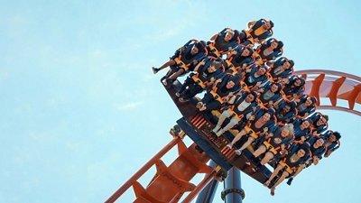 A roller coaster car full of excited riders, strapped in with orange harnesses, tilts downward against a clear blue sky.