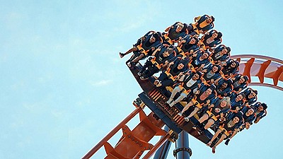 A roller coaster car full of excited riders, strapped in with orange harnesses, tilts downward against a clear blue sky.