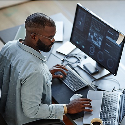  Man analyzing data on a monitor and laptop at his desk.