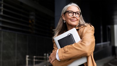 A smiling older woman with long gray hair and glasses stands outdoors, wearing a tan leather jacket and hugging a laptop and notebook to her chest while looking up and to the side.