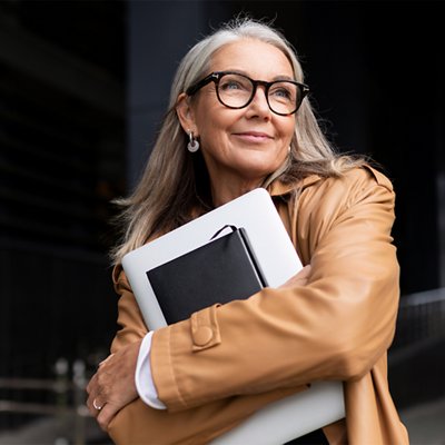 A smiling older woman with long gray hair and glasses stands outdoors, wearing a tan leather jacket and hugging a laptop and notebook to her chest while looking up and to the side.
