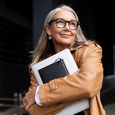 A smiling older woman with long gray hair and glasses stands outdoors, wearing a tan leather jacket and hugging a laptop and notebook to her chest while looking up and to the side.