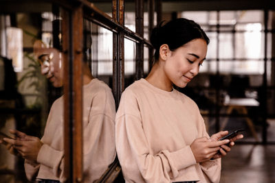 Image of young beautiful asian woman smiling and holding cellphone while working in office