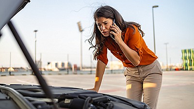 Woman on the phone looking under the hood of a car