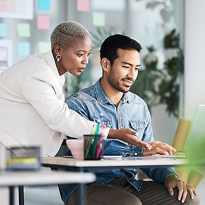 Two professionals collaborating at a computer in a modern office with sticky notes and plants in the background.