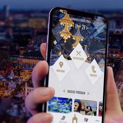 A hand holds a smartphone displaying an event or theme park app, with a blurred festive outdoor scene and lights in the background.