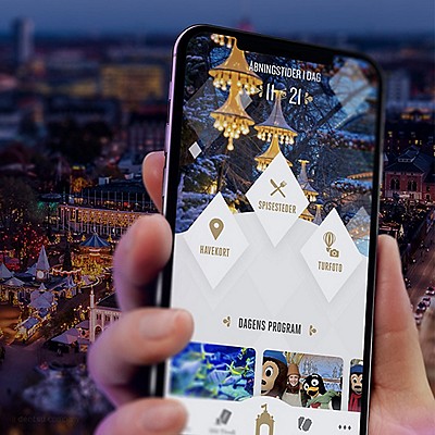 A hand holds a smartphone displaying an event or theme park app, with a blurred festive outdoor scene and lights in the background.