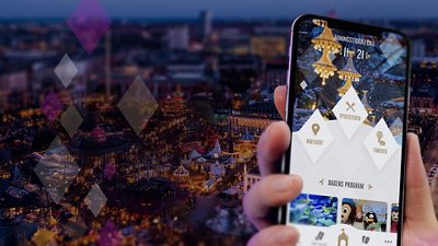 A hand holds a smartphone displaying an event or theme park app, with a blurred festive outdoor scene and lights in the background.