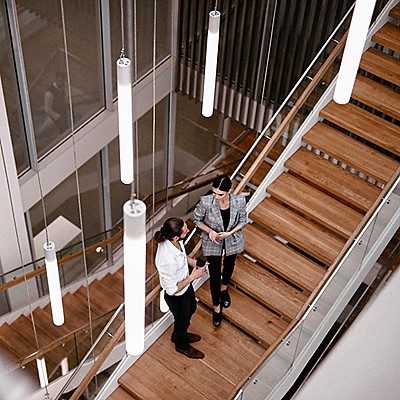 Two people standing and talking on a modern wooden staircase inside a contemporary office building with glass railings, large windows, and hanging cylindrical pendant lights.