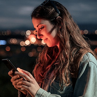 Person with long hair wearing a denim jacket and headphones, looking at a smartphone in a blurred evening setting with bokeh lights.