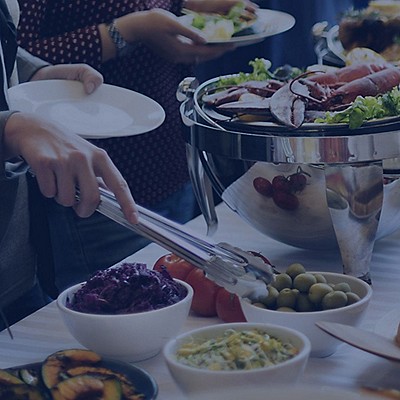 A hand with a spoon serving food onto a plate from a full table