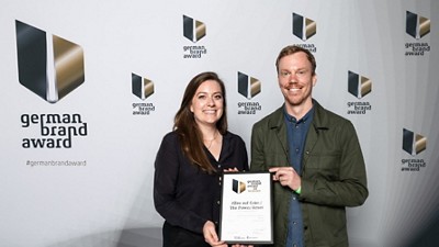 Ein Mann und eine Frau stehen zusammen und halten eine Urkunde bei der Veranstaltung des German Brand Award, im Hintergrund sind Markenlogos zu sehen.