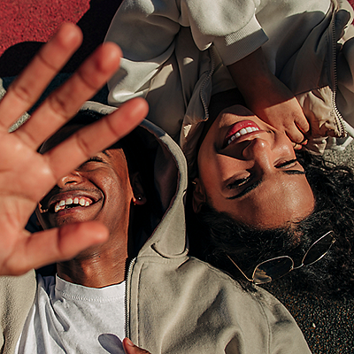 Two people lying on the ground, both smiling; one covers part of the camera with a hand, while the other faces up with sunglasses on their head.