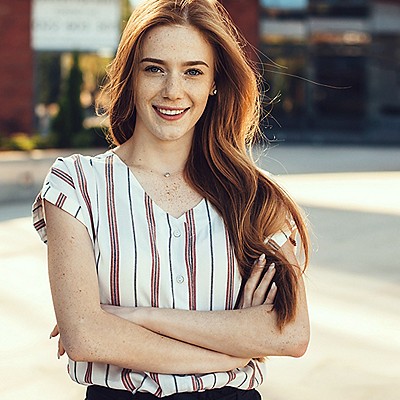 A young smiling woman with long hair and crossed arms stands facing forward.